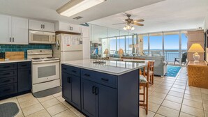 A modern kitchen with a central island, white cabinets, a microwave, and a stove. There is a dining area with a table and chairs, and a view of the ocean through large windows. - Oceanfront 3bed/2.5bath-Water's Edge Resort-612 Indoor/Outdoor Pool & Hot Tub (Garden City Beach)