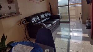A living room with a black leather sofa, a dining table with a blue table runner, a potted plant, and a sliding glass door leading to a balcony. - Rua Maria Tognini 450 (Praia Grande)