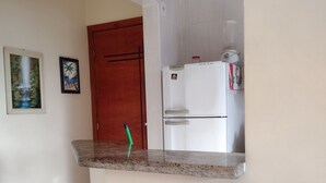 A kitchen with a white refrigerator, a wooden door, and a small table with a decorative item. - Rua Maria Tognini 450 (Praia Grande)
