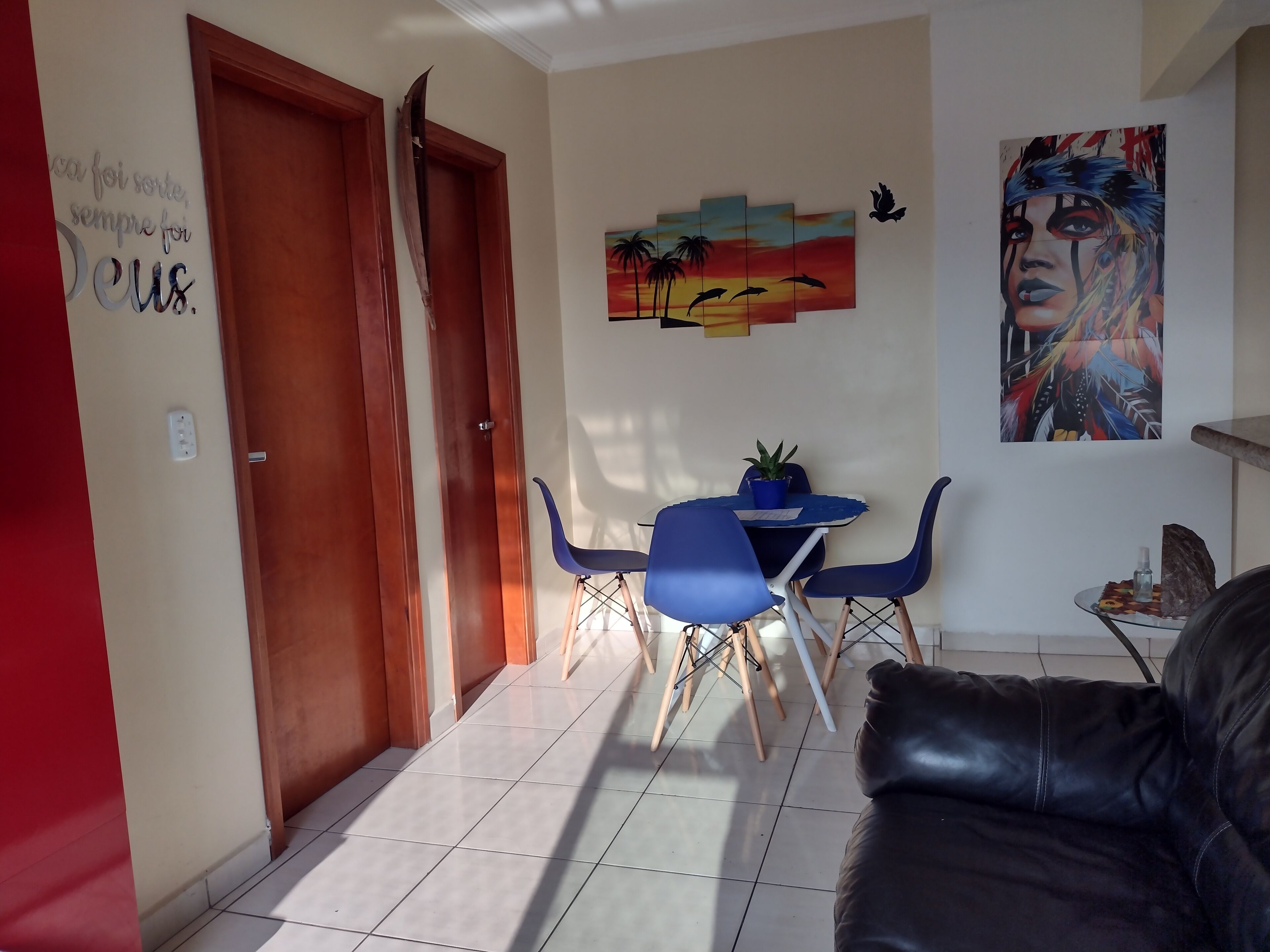 A living room with a black leather sofa, a dining area with blue chairs and a small table, a painting of a sunset, and a door with a wooden frame.