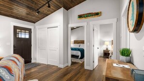 A spacious room with wooden flooring, a bed with a blue blanket, a desk with a lamp, and a door leading to another room.