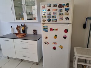A kitchen with a white refrigerator adorned with magnets, a glass cabinet with dishes, and a countertop with a basket and utensils. - Maison à Louer à 500 Mètres à Pieds de la Plage (LES ANSES D 'ARLET)