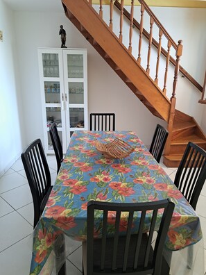 A dining area with a table covered in a floral tablecloth, surrounded by black chairs, a wooden staircase, and a cabinet with glass doors. - Maison à Louer à 500 Mètres à Pieds de la Plage (LES ANSES D 'ARLET)