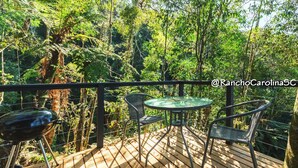 A wooden deck with a glass table and two chairs, surrounded by lush greenery and trees. - Rancho Carolina (Rancho Queimado)