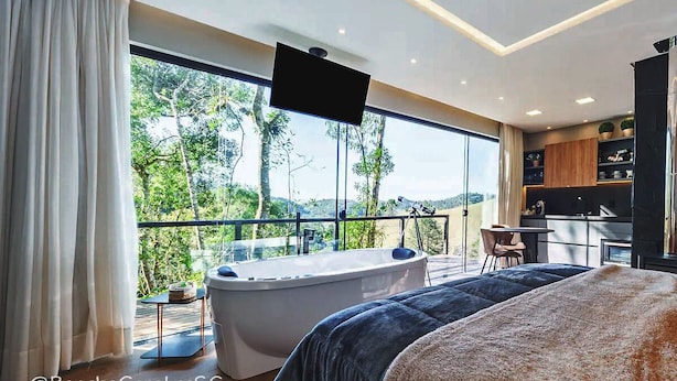 A modern bedroom with a large window, a bathtub, and a view of greenery. - Rancho Carolina (Rancho Queimado)