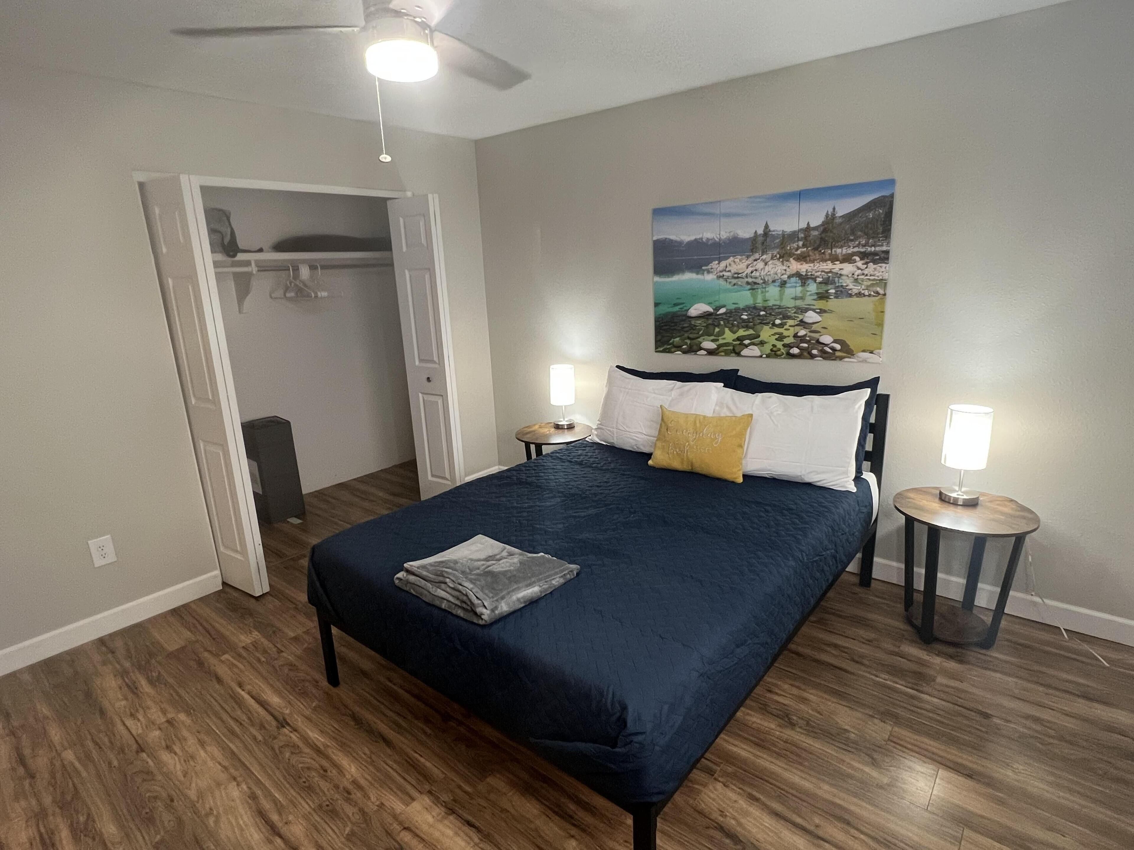 A bedroom with a bed, a painting of a lakeside scene, a closet, and two bedside lamps.