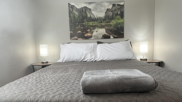 Een net opgemaakt bed met een gewatteerde deken, twee bedlampen en een lijst met een landschapsfoto erboven.