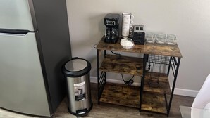 Een keuken met een roestvrijstalen koelkast, een koffieapparaat, een broodrooster en een prullenbak. Er staat een houten tafel onder een bord met koffiethema aan de muur.