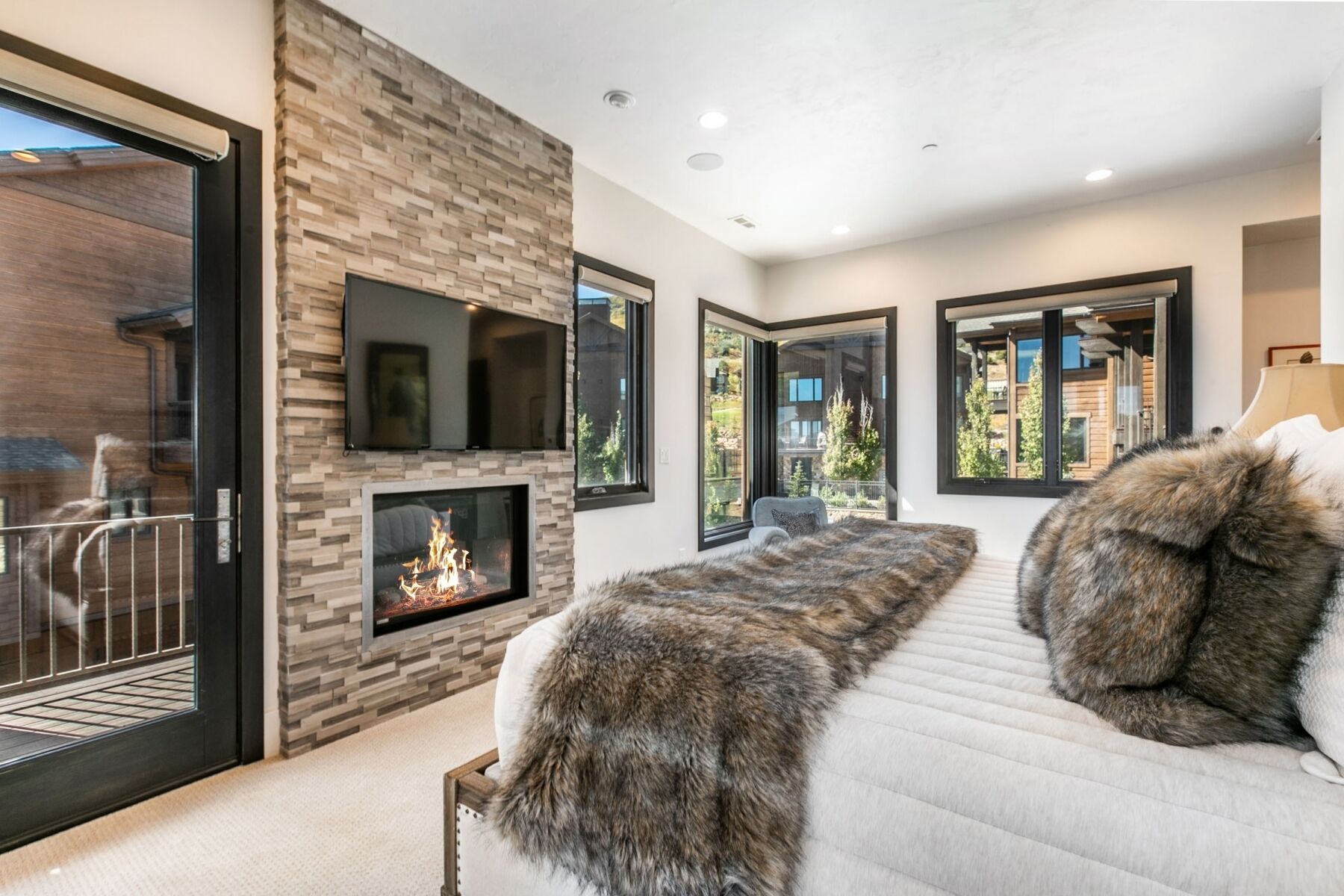 A modern living room with a fireplace, large windows, and a sofa with fur throw pillows.