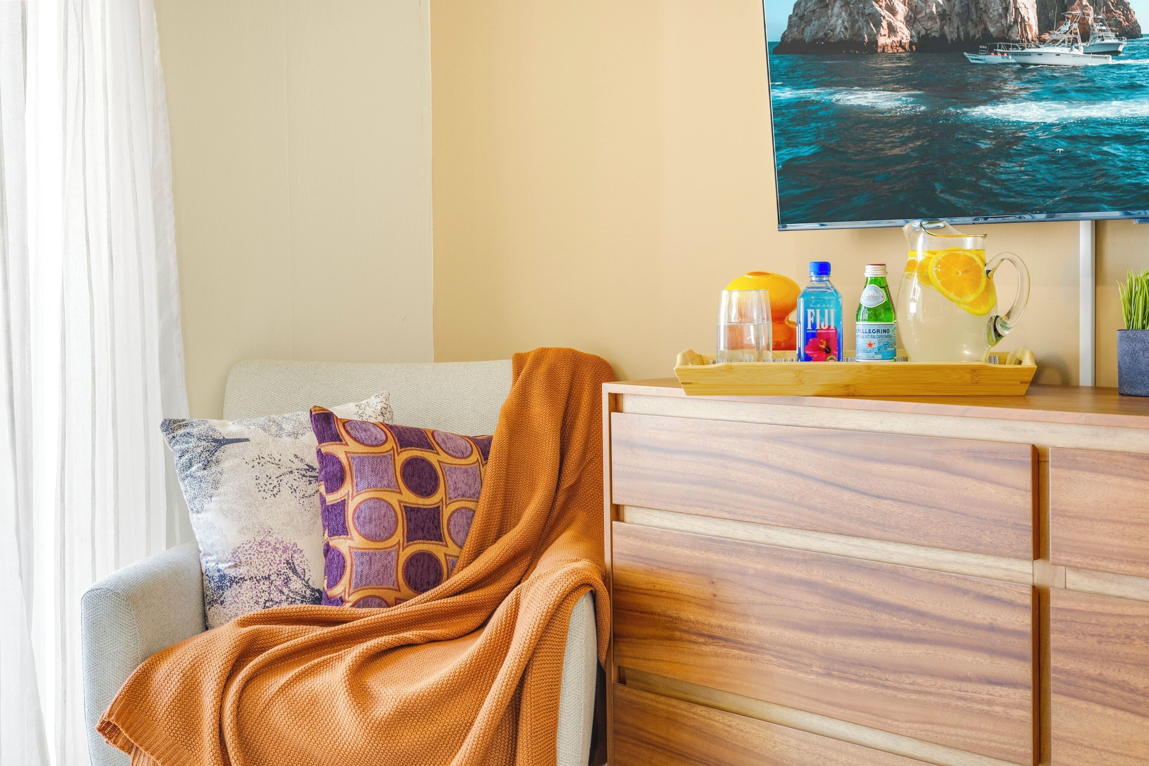 A living room with a sofa, a wooden cabinet, a television displaying a coastal scene, and a tray with beverages and a fruit.