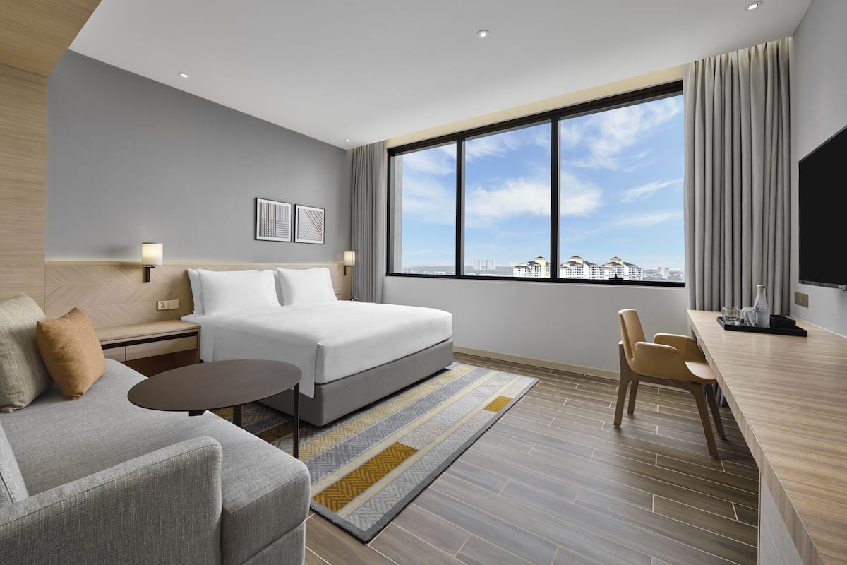A hotel room with a bed, a sofa, a small table, a TV, and a large window with a city view.