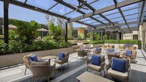 A covered outdoor seating area with wicker furniture and blue cushions, surrounded by greenery and a glass ceiling.