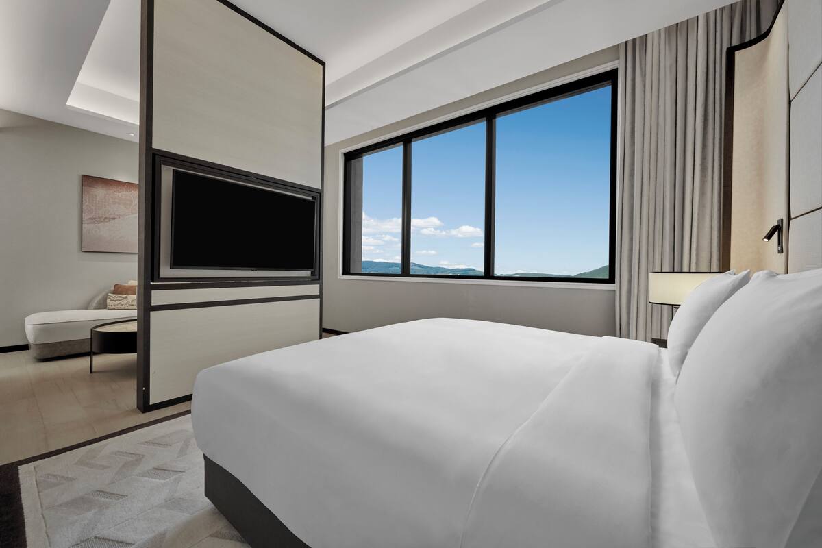 A modern hotel room with a large bed, a flat-screen TV, a sofa, and a large window offering a view of the mountains.