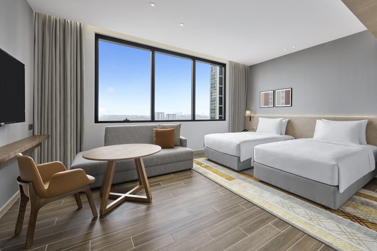 A modern hotel room with a large window, a sofa, two single beds, a small table, and a nightstand with a lamp.
