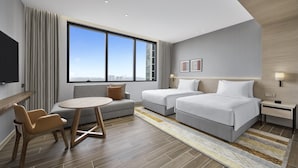 A modern hotel room with a large window, a sofa, two single beds, a small table, and a nightstand with a lamp.
