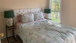 Un dormitorio con cabecera capitonada, ropa de cama floral, dos lámparas de noche, una ventana con vistas y un equipo de aire acondicionado.