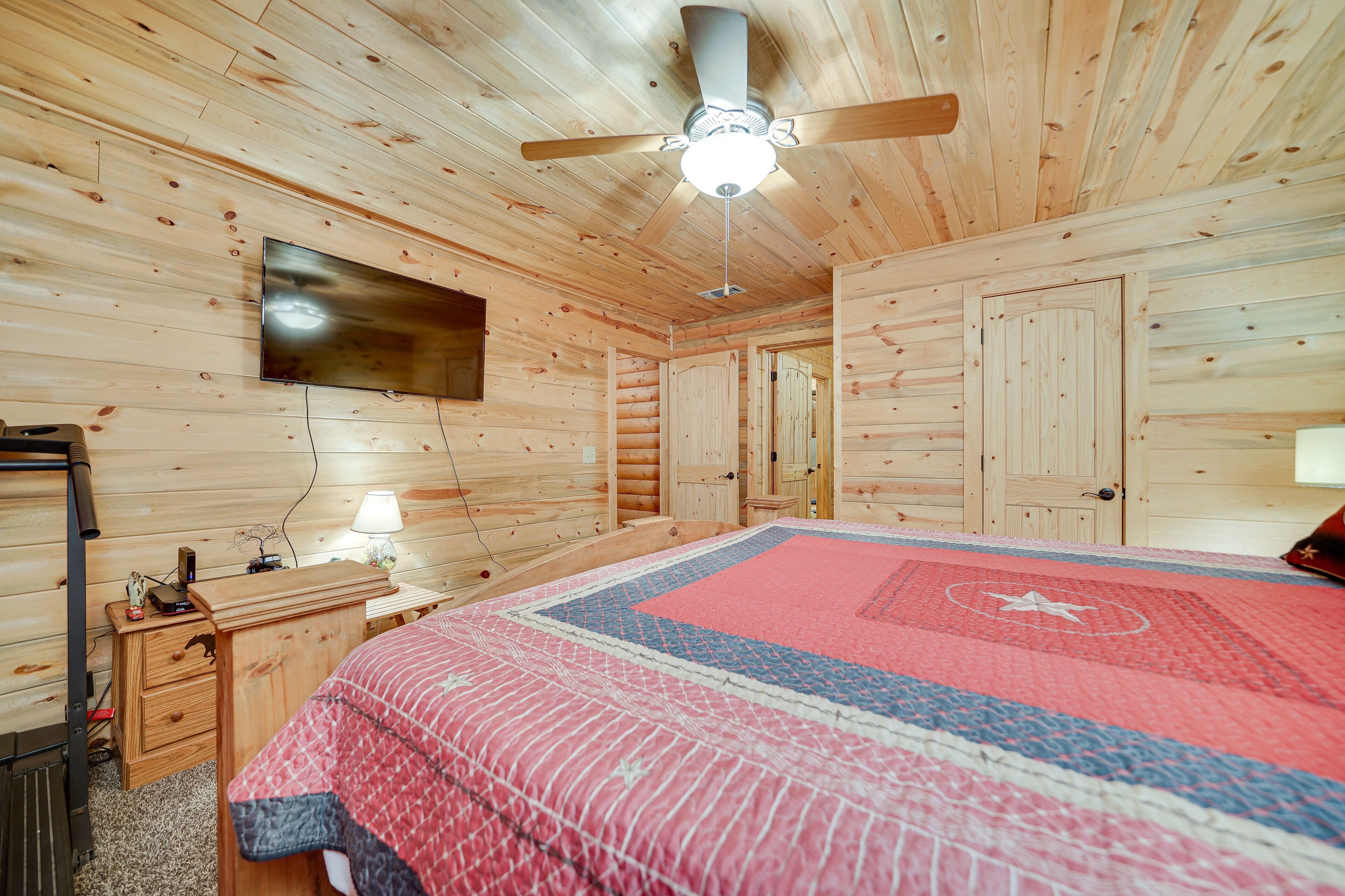 Quarto de cabana de madeira com cama, ventoinha no teto, televisão presa na parede e cómoda de madeira.