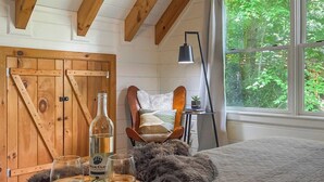 A cozy bedroom with a wooden ceiling, a large window with a view of greenery, a chair with a cushion, a fur throw, and a bottle of wine. - Leaf & Log Serene Cabin w/ Lounge Deck & Firepit (Cherry Log)