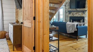 A cozy room with wooden walls, a stone fireplace, a blue sofa, and a television mounted on the wall. - Leaf & Log Serene Cabin w/ Lounge Deck & Firepit (Cherry Log)