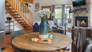 A cozy living room with a wooden staircase, a fireplace, and a round dining table with a plate of snacks. - Leaf & Log Serene Cabin w/ Lounge Deck & Firepit (Cherry Log)