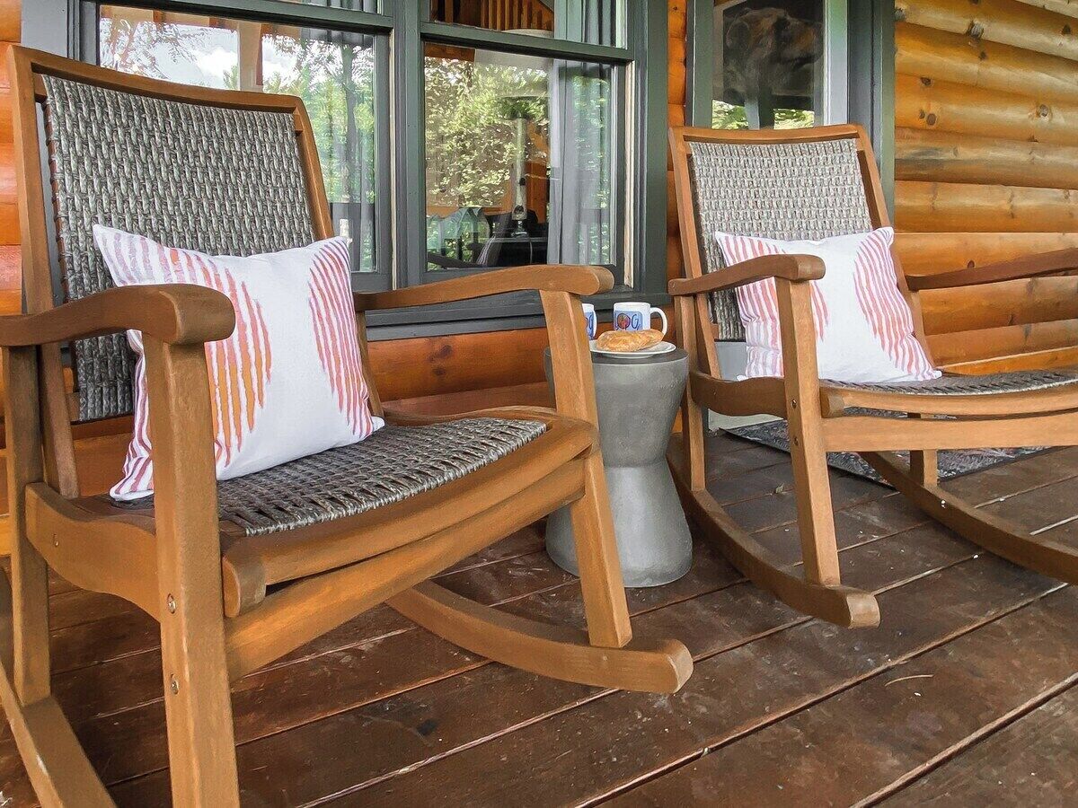 Dos mecedoras con asientos de mimbre y estructura de madera, ubicadas en una terraza de madera con una mesa de vidrio en el centro.