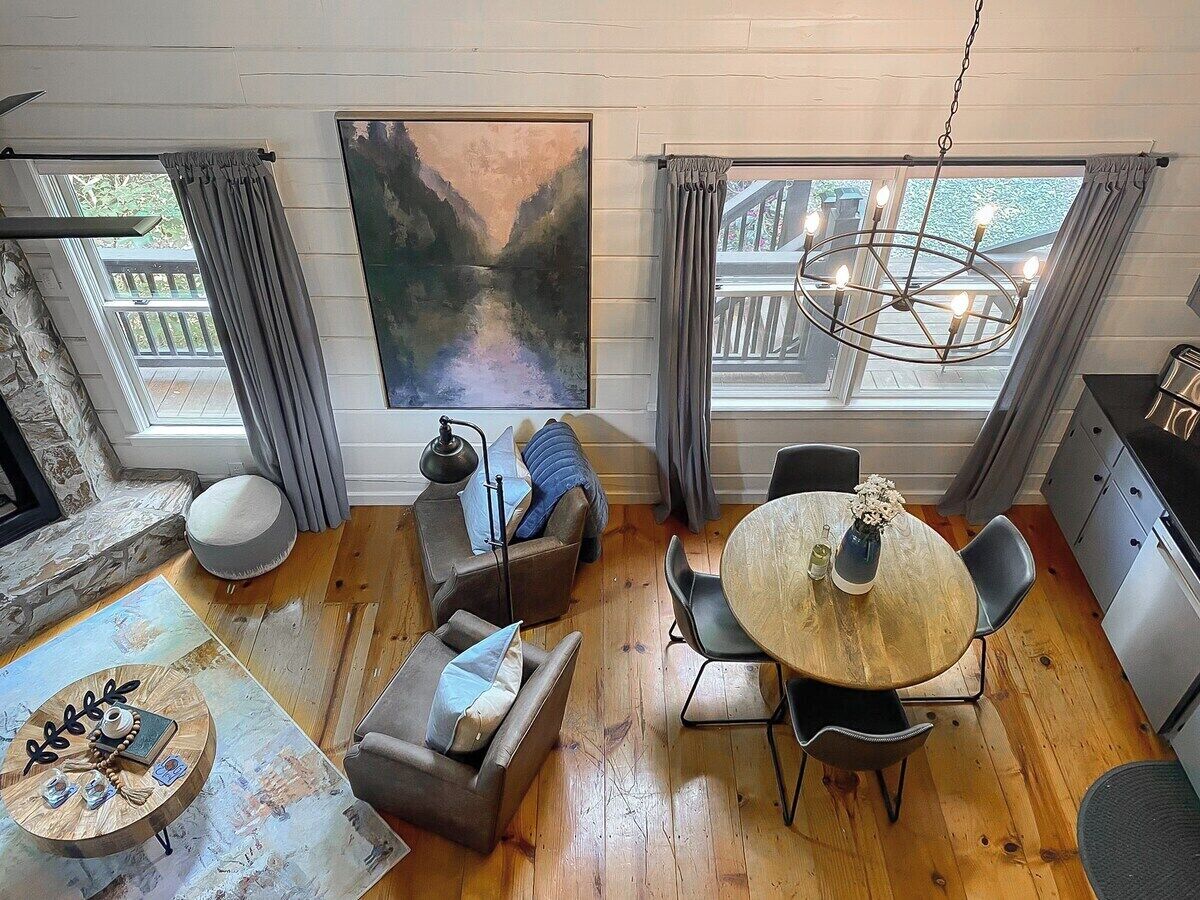 A cozy living room with a round wooden table, comfortable seating, a painting on the wall, and a view of a deck through large windows.