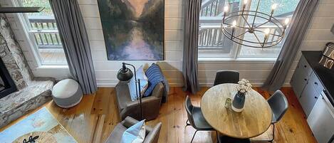 A cozy living room with a round wooden table, comfortable seating, a painting on the wall, and a view of a deck through large windows.