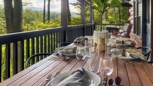 A wooden dining table set for a meal with plates, cutlery, and glasses, overlooking a lush landscape. - Leaf & Log Serene Cabin w/ Lounge Deck & Firepit (Cherry Log)