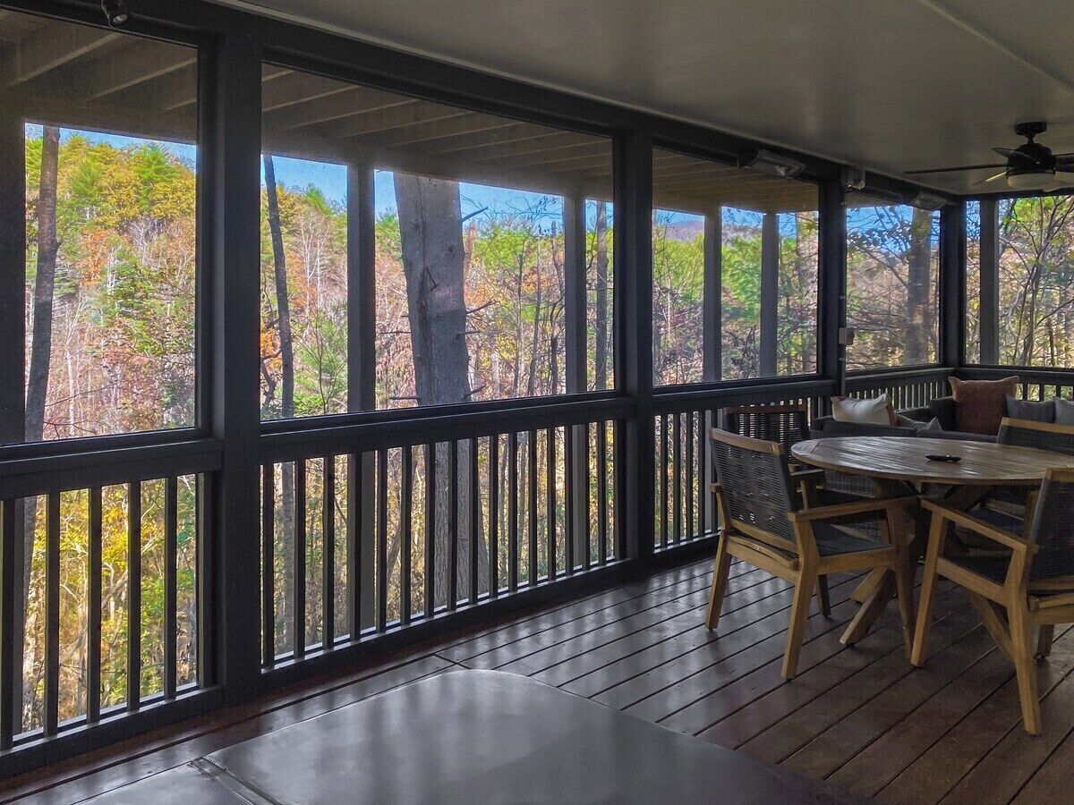 A covered outdoor area with a wooden table and chairs, a railing, and a view of trees.