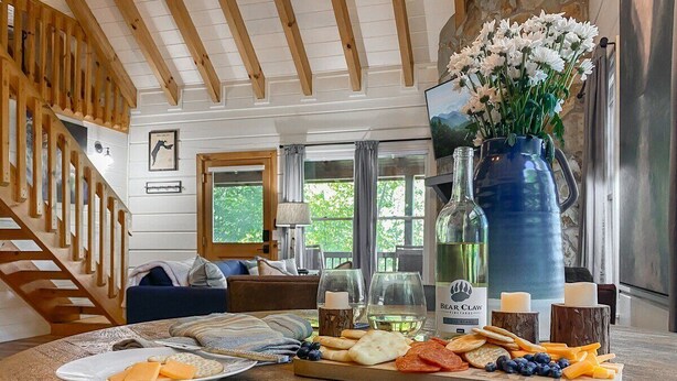 A wooden dining table with a cheese platter, wine bottle, and candles. A staircase and a living room are visible in the background. - Leaf & Log Serene Cabin w/ Lounge Deck & Firepit (Cherry Log)