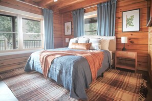 Un dormitorio con cama, mesita de noche, ventana e imagen colgada en la pared.