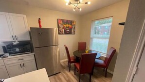 A compact kitchen with a refrigerator, microwave, and wooden cabinets. There is a dining area with a table and chairs, and a window with blinds. - Right by AT&T, Rangers stadium (Arlington)
