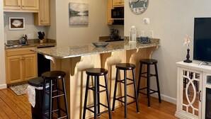 A kitchen with wooden cabinets, a granite countertop, a fish-themed clock, and a bar area with stools. - Cute waterfront bungalow, tons of amenities, a small gated fishing community! (Dauphin Island)