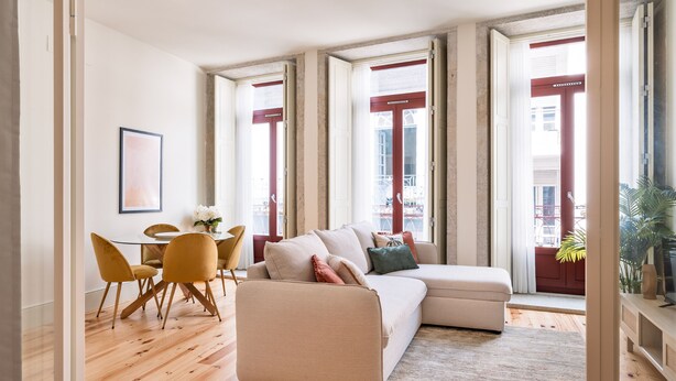 Un salon moderne avec un canapé beige, des fauteuils jaunes, une table à manger et de grandes fenêtres aux cadres rouges.