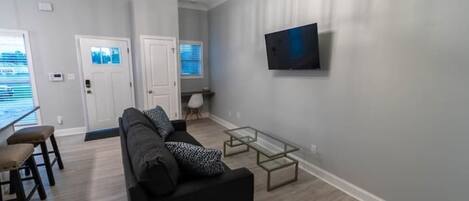 Ein modernes Wohnzimmer mit einer schwarzen Couch, einem Glastisch und einem an der Wand befestigten Fernseher.