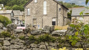 A stone building with a gabled roof, a central vertical window, and a stone wall in the foreground. - Church View (Grange-over-Sands)