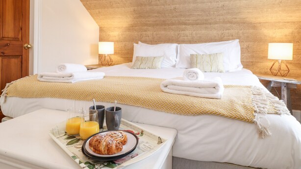 A hotel room with a wooden ceiling, a bed with white linens, two bedside lamps, and a breakfast tray with croissant and orange juice. - Church View (Grange-over-Sands)