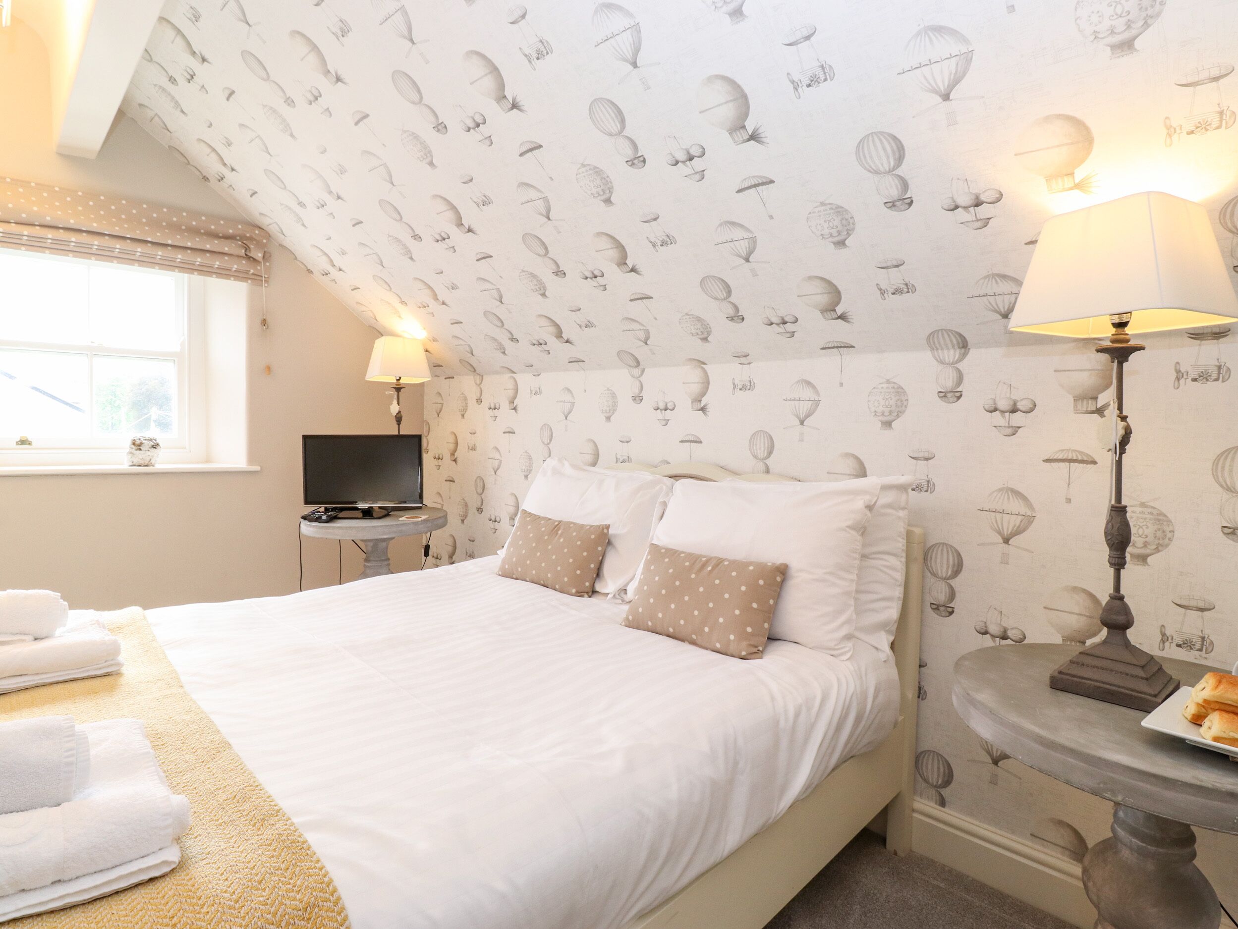 A bedroom with a bed, pillows, a bedside table, a lamp, a TV, and a window.
