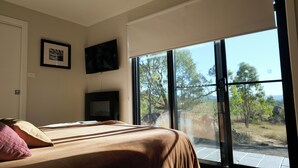 A bedroom with a large window, a bed, a flat-screen TV, and framed artwork on the wall. - Golden Hour Views: Sunset Cabin with Outdoor Bath (Frog Rock)
