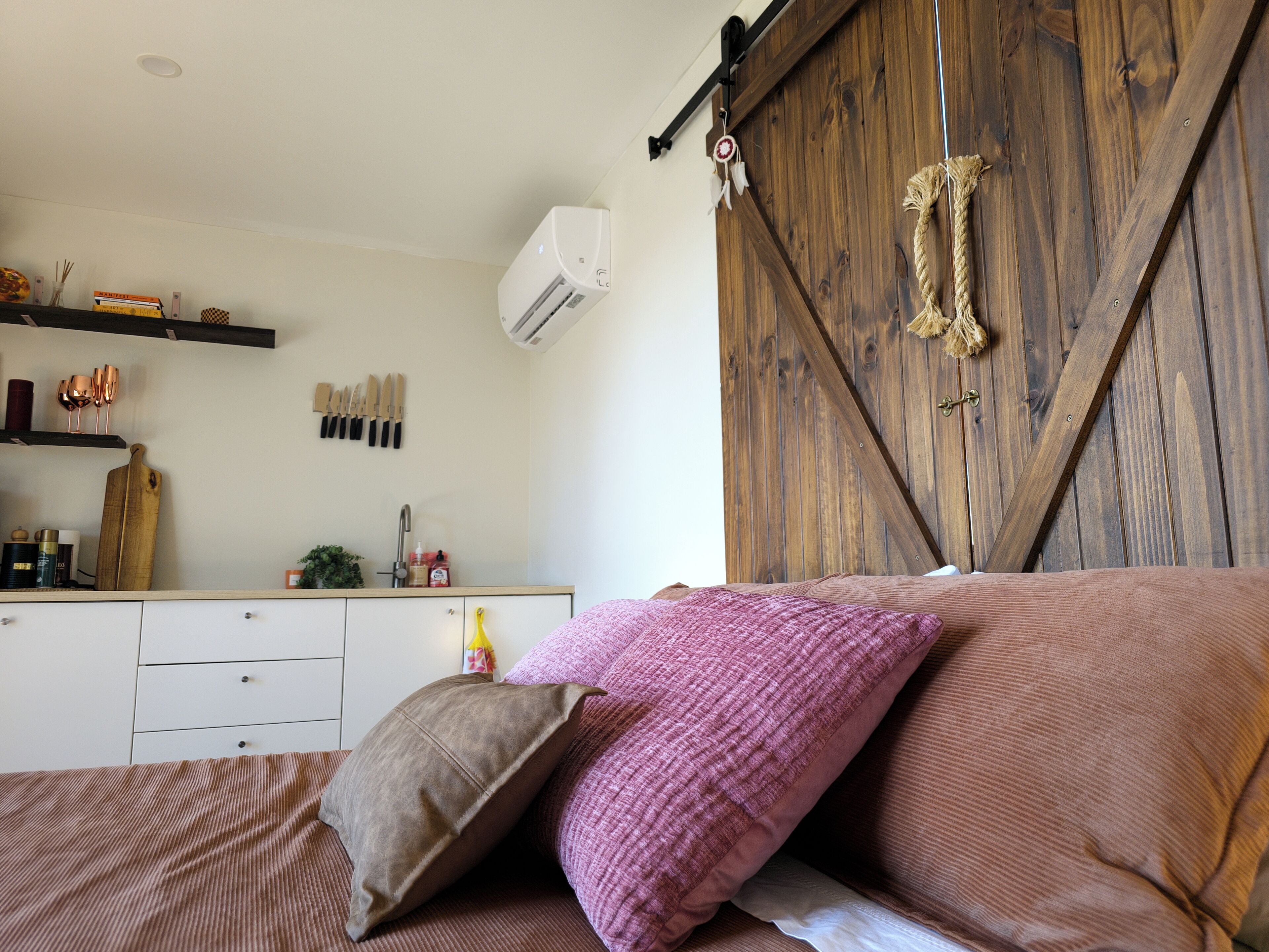 Un dormitorio con cama, armario de madera, área de cocina con utensilios y aire acondicionado de pared.