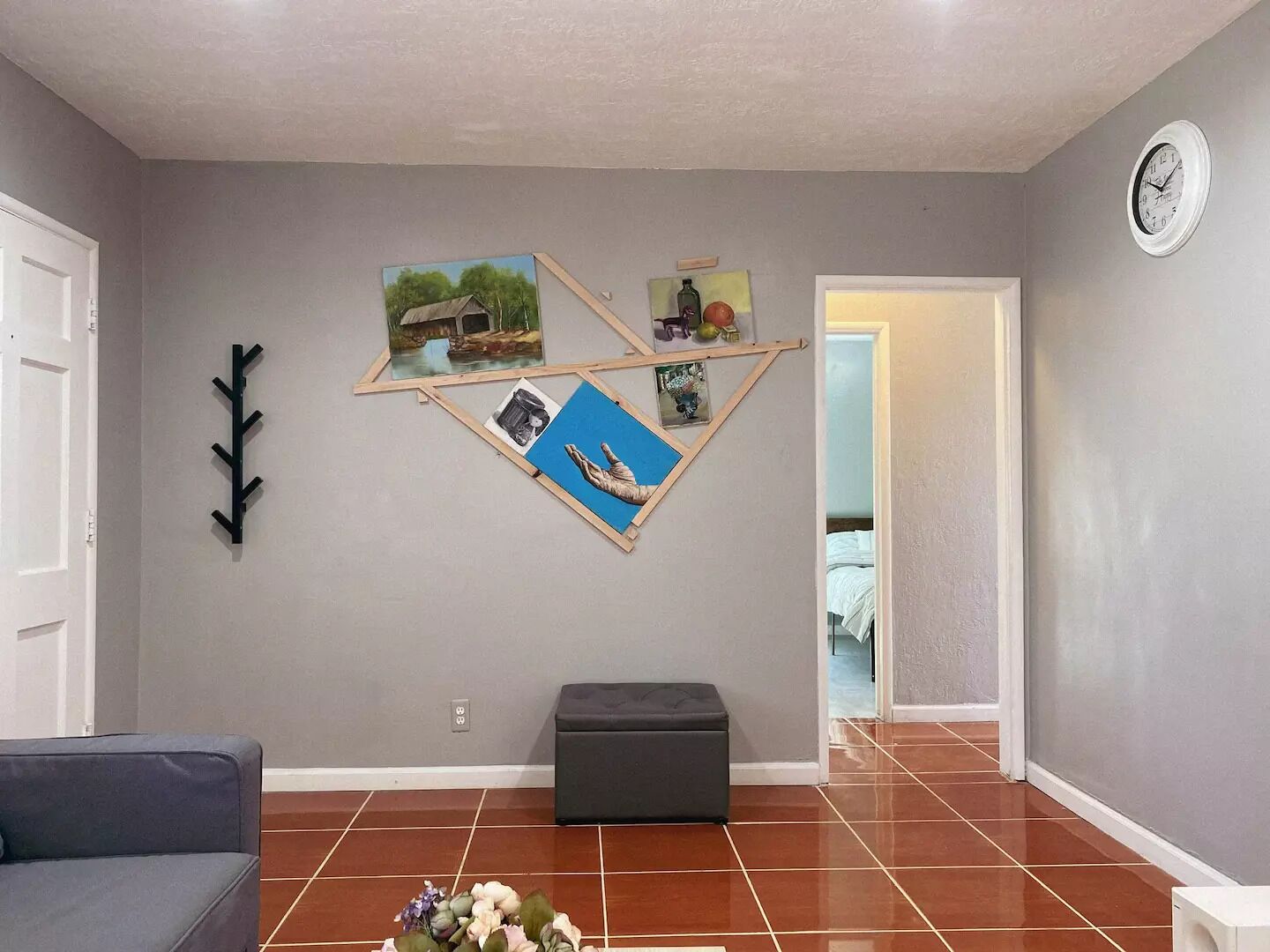 Un salon avec un mur gris, un plancher en bois, un canapé gris, un pouf gris et une horloge au mur.