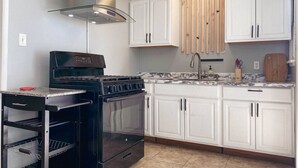Une cuisine avec des meubles blancs, une cuisinière noire et une étagère en bois au-dessus de l’évier.