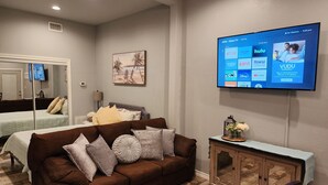 Ein Wohnzimmer mit braunem Sofa, einem an der Wand montierten Fernseher, der Streaming-Dienste anzeigt, einem kleinen Schrank und einem Bild an der Wand.