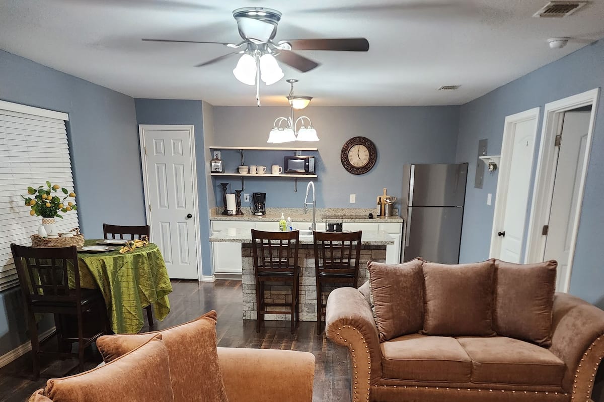 A living room with two brown sofas, a dining table with chairs, a kitchen area with a fridge, microwave, and sink, and a ceiling fan.