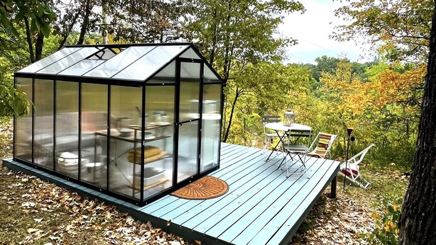 A glass greenhouse structure with a wooden deck and a table with chairs. - Cedar Springs Greenhouse (Decorah)