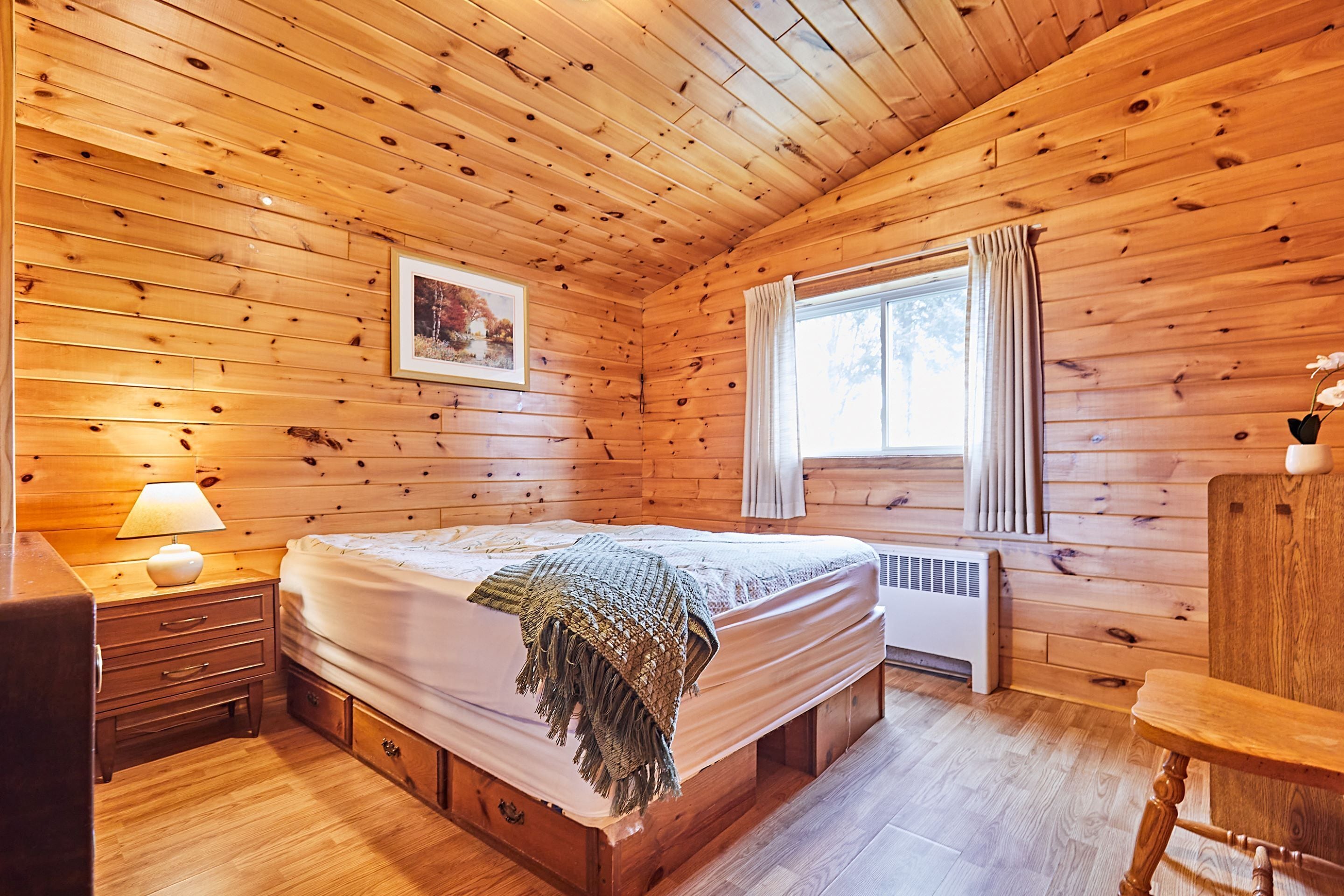 Quarto de cabana de madeira com cama, mesa de cabeceira, abajur e janela com cortinas.