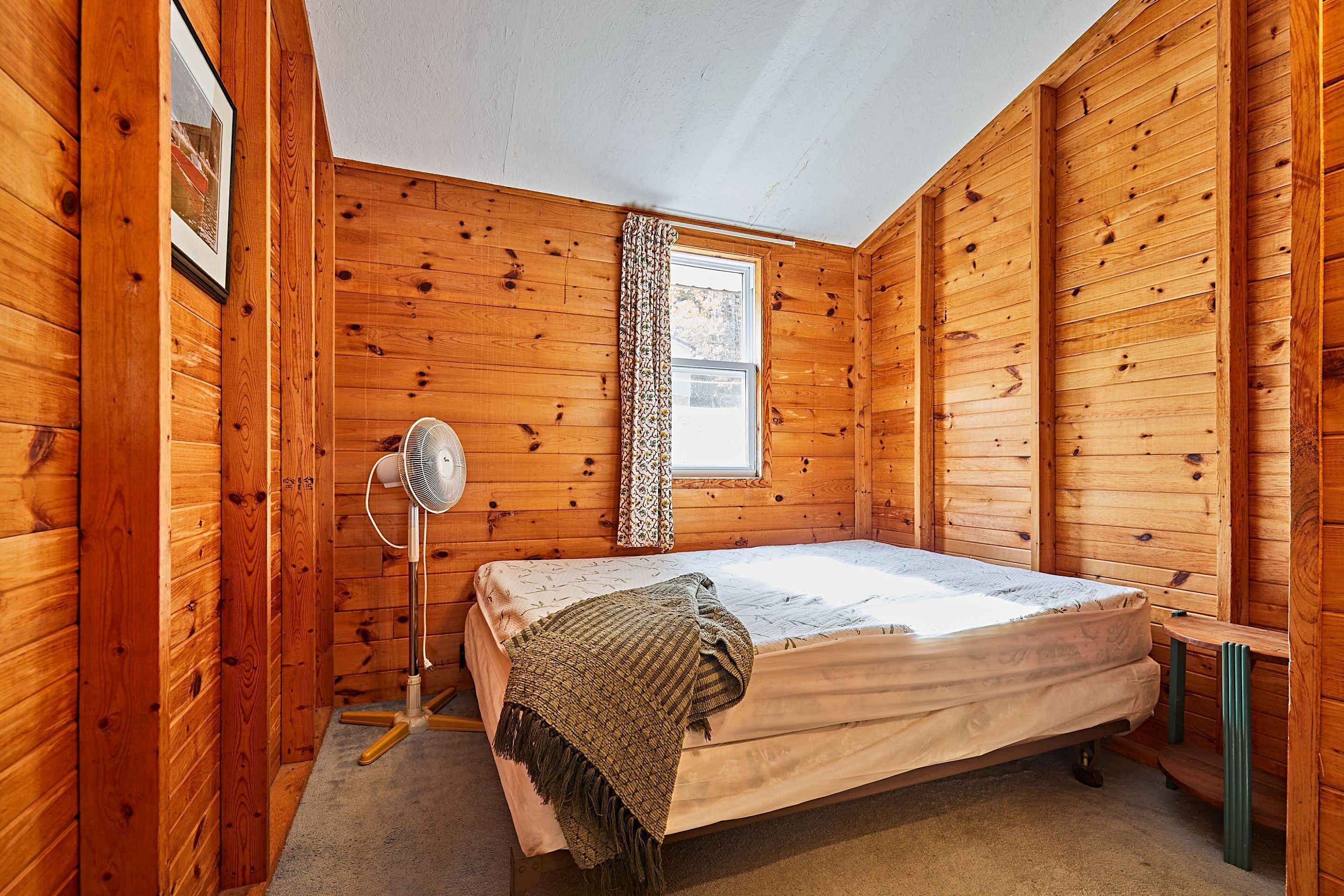 A wooden cabin room with a bed, a standing fan, and a window with curtains.