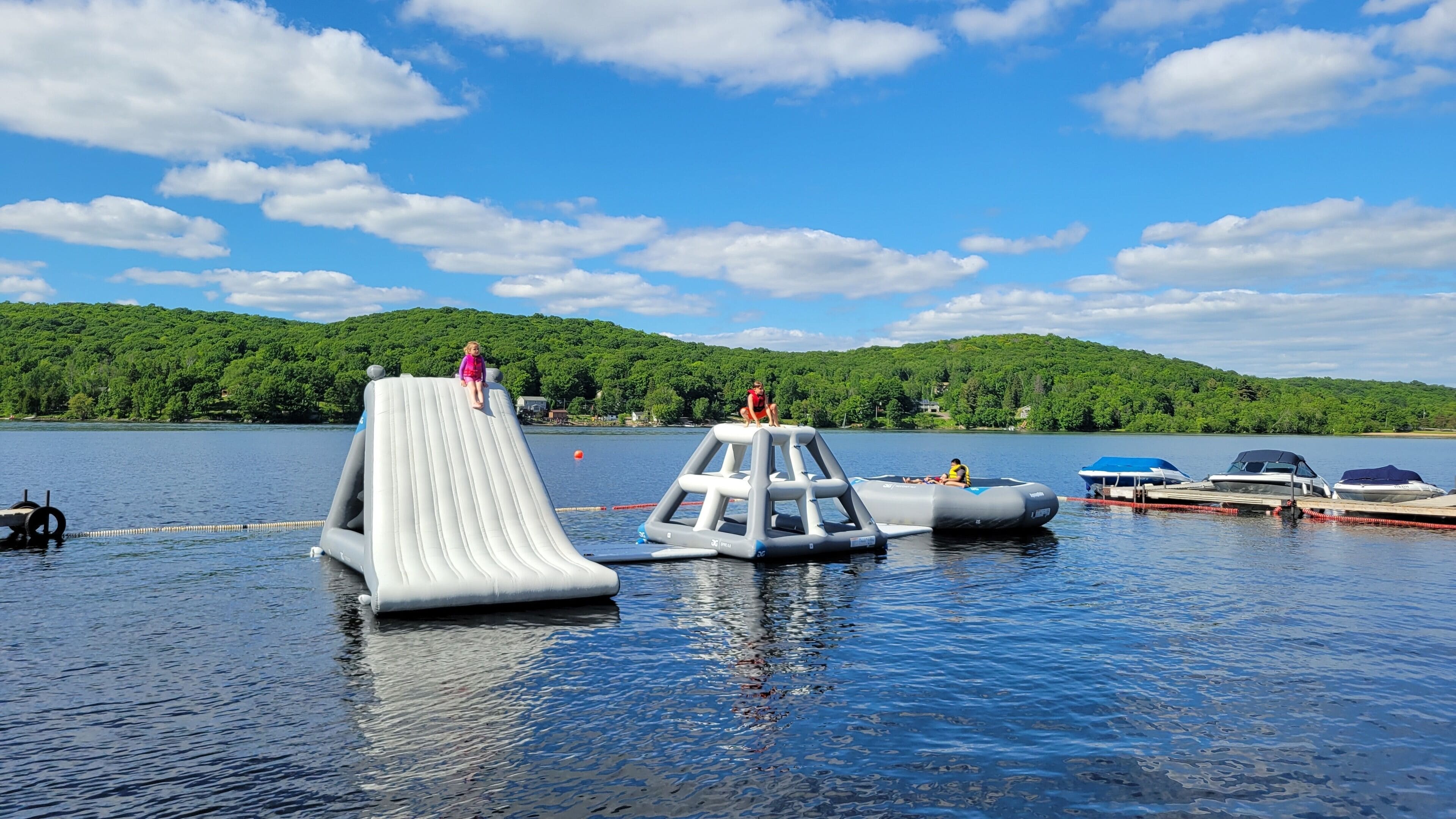 An inflatable water park with a slide and play structures on a lake, surrounded by boats and a forested hillside.