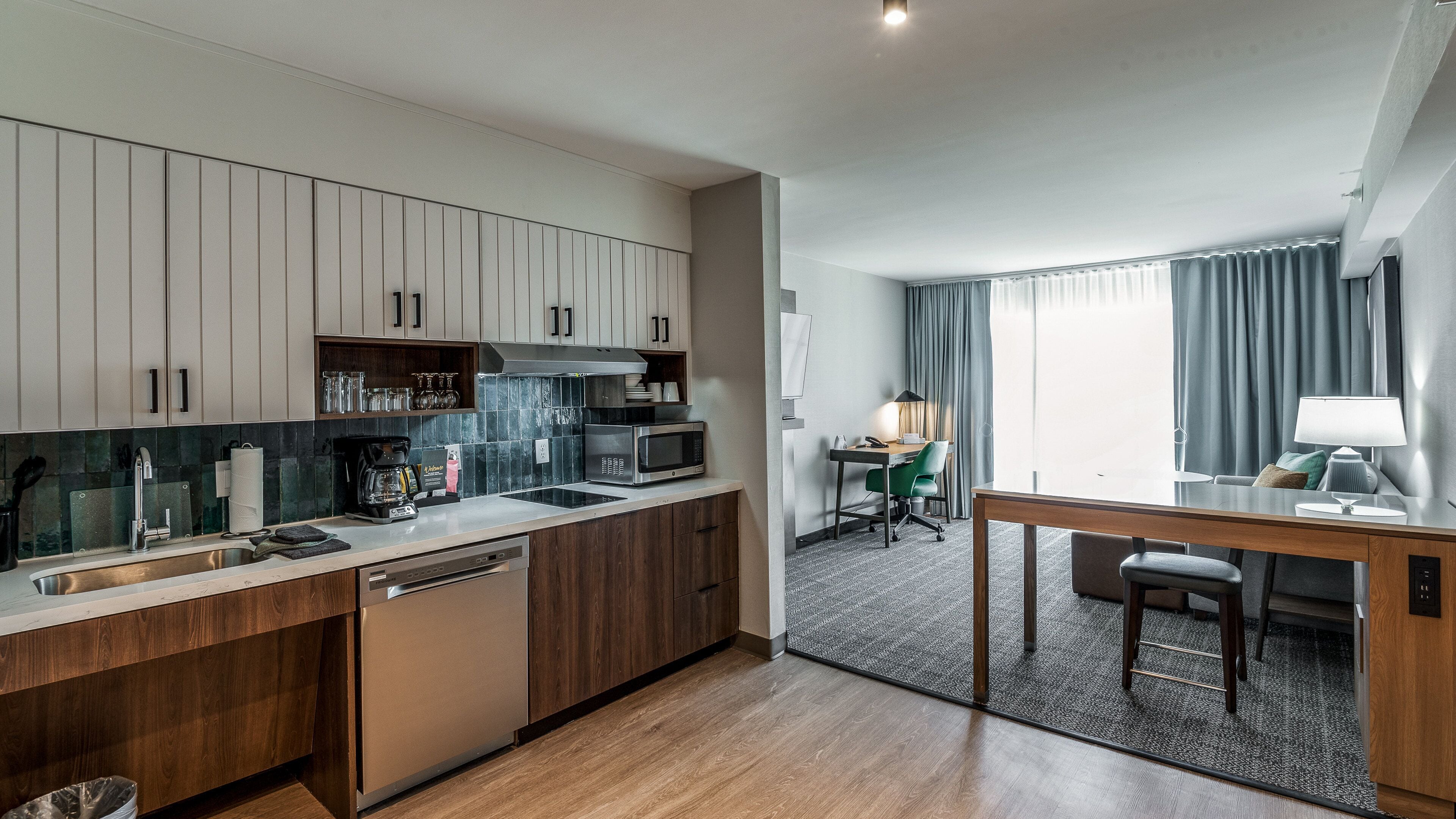 A modern kitchen with wooden cabinets, a built-in microwave, and a coffee maker. There is a dining area with a table and chairs, and a window with curtains.