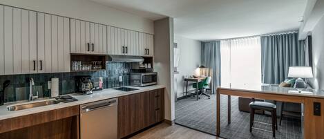 A modern kitchen with wooden cabinets, a built-in microwave, and a coffee maker. There is a dining area with a table and chairs, and a window with curtains.